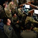 A group of military personnel from multiple branches and possibly allied nations are gathered in what appears to be a command operations center or joint operations facility. Personnel are wearing various uniforms including US Army/Air Force OCP camouflage, Navy digital camouflage, and olive/sage flight suits with rank insignia and unit patches. The scene suggests a briefing, training exercise, or joint operational planning session at workstations equipped with multiple computer monitors.