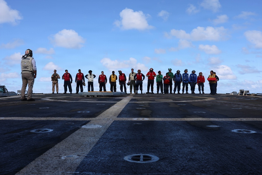 A person with 'BMC' visible on their back stands facing a line of approximately 20 personnel on what appears to be a naval ship's flight deck or helicopter landing pad. The personnel are wearing various colored safety vests (red, yellow, green, blue, white) and appear to be assembled for a briefing or muster. The setting is an open ocean environment under a partly cloudy sky.