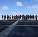 A person with 'BMC' visible on their back stands facing a line of approximately 20 personnel on what appears to be a naval ship's flight deck or helicopter landing pad. The personnel are wearing various colored safety vests (red, yellow, green, blue, white) and appear to be assembled for a briefing or muster. The setting is an open ocean environment under a partly cloudy sky.