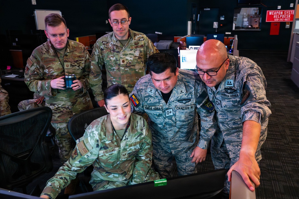 A group of five military personnel, including U.S. Air Force members and what appear to be South American air force officers (identified by 'Fuerza Aerea' patches), are gathered around a computer workstation in what appears to be a military operations or training facility. The personnel are collaborating and reviewing content on a monitor, suggesting a joint training or briefing session. Name tapes visible include 'Anselmo,' 'Cantos R.,' and partially visible others.