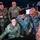 A group of five military personnel, including U.S. Air Force members and what appear to be South American air force officers (identified by 'Fuerza Aerea' patches), are gathered around a computer workstation in what appears to be a military operations or training facility. The personnel are collaborating and reviewing content on a monitor, suggesting a joint training or briefing session. Name tapes visible include 'Anselmo,' 'Cantos R.,' and partially visible others.