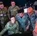 A group of five military personnel, including U.S. Air Force members and what appear to be South American air force officers (identified by 'Fuerza Aerea' patches), are gathered around a computer workstation in what appears to be a military operations or training facility. The personnel are collaborating and reviewing content on a monitor, suggesting a joint training or briefing session. Name tapes visible include 'Anselmo,' 'Cantos R.,' and partially visible others.