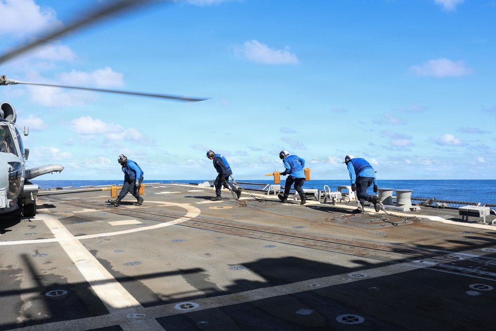Four naval personnel in blue uniforms are working on the flight deck of a military vessel at sea, handling chains and tie-down equipment near a helicopter. The crew members appear to be securing or releasing aircraft tie-down chains on what appears to be a destroyer or frigate flight deck. The operation is taking place under clear blue skies with calm ocean conditions visible in the background.