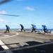 Four naval personnel in blue uniforms are working on the flight deck of a military vessel at sea, handling chains and tie-down equipment near a helicopter. The crew members appear to be securing or releasing aircraft tie-down chains on what appears to be a destroyer or frigate flight deck. The operation is taking place under clear blue skies with calm ocean conditions visible in the background.