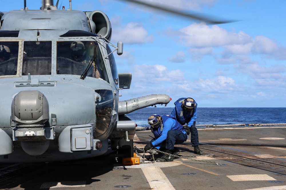 Two naval personnel in blue flight deck suits and helmets are securing a military helicopter to a ship's flight deck using chains and tie-down equipment. The helicopter, likely an MH-60 Seahawk variant, is positioned on the deck of a naval vessel at sea. The operation appears to be a standard chock-and-chain securing procedure following a helicopter landing.