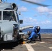 Two naval personnel in blue flight deck suits and helmets are securing a military helicopter to a ship's flight deck using chains and tie-down equipment. The helicopter, likely an MH-60 Seahawk variant, is positioned on the deck of a naval vessel at sea. The operation appears to be a standard chock-and-chain securing procedure following a helicopter landing.