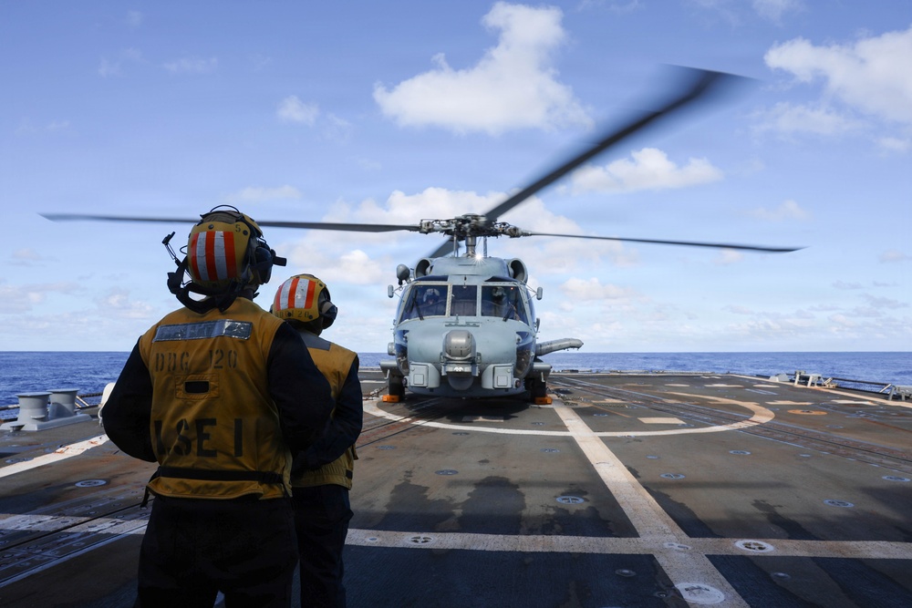 Two naval personnel in yellow safety vests and helmets stand on the flight deck of a U.S. Navy destroyer, observing a helicopter with spinning rotors on the landing pad. The vest of the taller individual is marked 'DDG 120' and 'LSE 1', identifying the ship and the role of Landing Signal Enlisted. The scene takes place at sea under partly cloudy skies.
