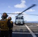 Two naval personnel in yellow safety vests and helmets stand on the flight deck of a U.S. Navy destroyer, observing a helicopter with spinning rotors on the landing pad. The vest of the taller individual is marked 'DDG 120' and 'LSE 1', identifying the ship and the role of Landing Signal Enlisted. The scene takes place at sea under partly cloudy skies.