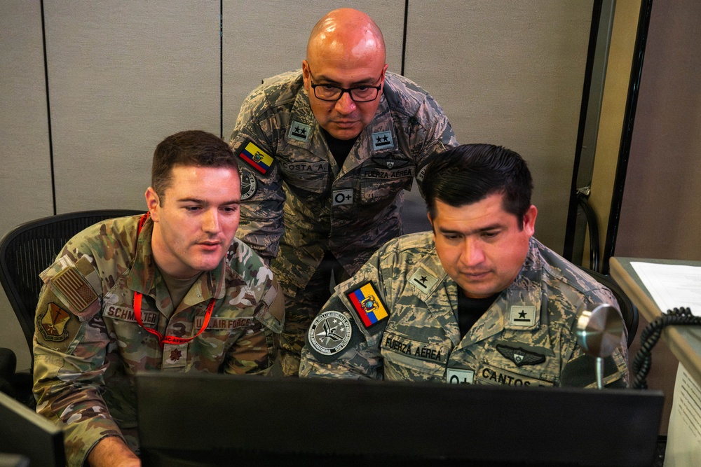 Three military personnel from different nations are gathered around a computer monitor in what appears to be a command or operations center. Two individuals wear Ecuadorian Air Force ('Fuerza Aerea') uniforms with Ecuadorian flag patches, while one wears a U.S. Air Force multicam uniform. They appear to be engaged in a joint military exercise or collaborative work session.