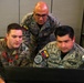 Three military personnel from different nations are gathered around a computer monitor in what appears to be a command or operations center. Two individuals wear Ecuadorian Air Force ('Fuerza Aerea') uniforms with Ecuadorian flag patches, while one wears a U.S. Air Force multicam uniform. They appear to be engaged in a joint military exercise or collaborative work session.