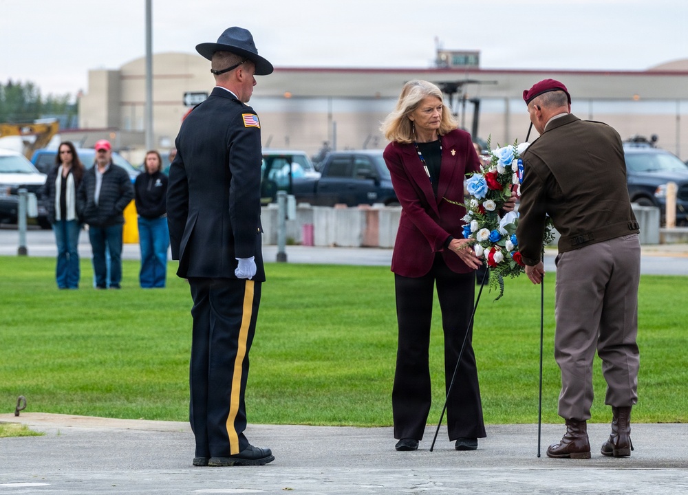 A formal wreath-laying ceremony is taking place outdoors, likely at a military installation or memorial site. A civilian woman in a burgundy blazer is participating alongside two military personnel, one in dress uniform with a campaign cover and the other in olive/tan combat uniform with a maroon beret. Bystanders observe in the background near a parking area.