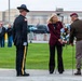 A formal wreath-laying ceremony is taking place outdoors, likely at a military installation or memorial site. A civilian woman in a burgundy blazer is participating alongside two military personnel, one in dress uniform with a campaign cover and the other in olive/tan combat uniform with a maroon beret. Bystanders observe in the background near a parking area.