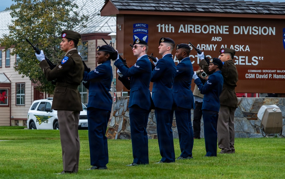 A military honor guard ceremony is taking place on a grassy area in front of a sign for the 11th Airborne Division and U.S. Army Alaska 'Arctic Angels.' Personnel in U.S. Air Force dress blue uniforms and at least one soldier in Army service uniform are performing a rifle salute or ceremonial drill. The sign also references Command Sergeant Major David P. Hanson, and a military police vehicle is visible in the background.