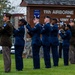 A military honor guard ceremony is taking place on a grassy area in front of a sign for the 11th Airborne Division and U.S. Army Alaska 'Arctic Angels.' Personnel in U.S. Air Force dress blue uniforms and at least one soldier in Army service uniform are performing a rifle salute or ceremonial drill. The sign also references Command Sergeant Major David P. Hanson, and a military police vehicle is visible in the background.