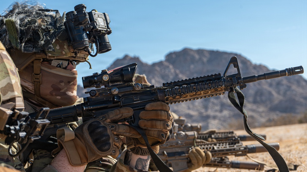 A soldier in full tactical gear is in a prone or low firing position in a desert environment, aiming an M4/M16-style carbine rifle. The soldier is equipped with advanced optics, night vision devices, and multicam camouflage. Additional armed personnel are visible in the background, suggesting a training exercise or operational deployment.