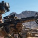 A soldier in full tactical gear is in a prone or low firing position in a desert environment, aiming an M4/M16-style carbine rifle. The soldier is equipped with advanced optics, night vision devices, and multicam camouflage. Additional armed personnel are visible in the background, suggesting a training exercise or operational deployment.
