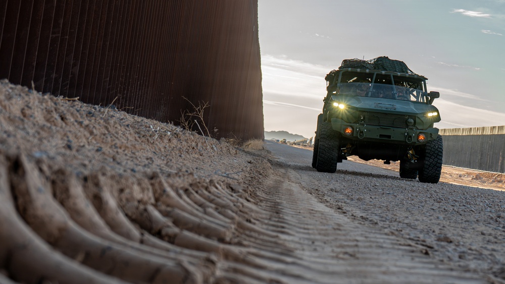 A military-style tactical vehicle is patrolling along a border wall on a dirt road at dusk or dawn. The vehicle, painted in olive drab/military green, is traveling parallel to a tall steel border barrier with its headlights illuminated. The foreground shows disturbed soil with visible root structures or debris along the base of the wall.