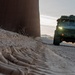 A military-style tactical vehicle is patrolling along a border wall on a dirt road at dusk or dawn. The vehicle, painted in olive drab/military green, is traveling parallel to a tall steel border barrier with its headlights illuminated. The foreground shows disturbed soil with visible root structures or debris along the base of the wall.