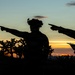 Two military personnel are silhouetted against a desert sunset sky, both pointing in the same direction while one holds what appears to be a map or document. The scene is set in an arid desert environment with Joshua trees and sparse vegetation visible in the background. The dramatic backlighting creates a high-contrast silhouette composition with warm orange and cool blue tones in the sky.