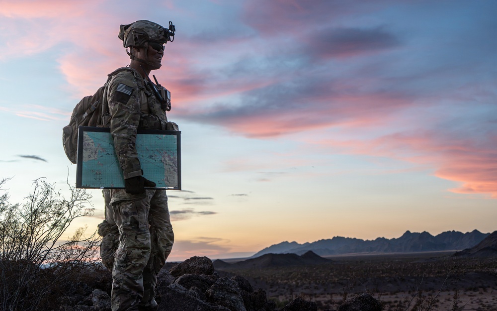 A U.S. Army soldier in full combat gear stands on elevated rocky terrain at dusk, holding a large laminated tactical map while surveying a desert landscape. The soldier is equipped with night vision device mount on helmet, a radio/communication device, tactical backpack, and an American flag patch visible on the left shoulder. The dramatic sunset sky with pink and blue hues frames the silhouetted mountain range in the background.