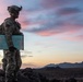 A U.S. Army soldier in full combat gear stands on elevated rocky terrain at dusk, holding a large laminated tactical map while surveying a desert landscape. The soldier is equipped with night vision device mount on helmet, a radio/communication device, tactical backpack, and an American flag patch visible on the left shoulder. The dramatic sunset sky with pink and blue hues frames the silhouetted mountain range in the background.