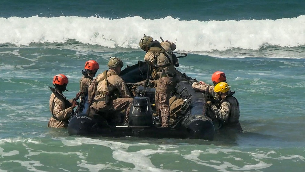 A group of military personnel in camouflage uniforms and colored helmets are maneuvering a black inflatable rubber combat raiding craft (CRRC) through ocean surf. The scene appears to depict a beach landing or surf passage training exercise, likely conducted by special operations or amphibious forces. The personnel are working collectively to control the craft against breaking waves.