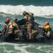 A group of military personnel in camouflage uniforms and colored helmets are maneuvering a black inflatable rubber combat raiding craft (CRRC) through ocean surf. The scene appears to depict a beach landing or surf passage training exercise, likely conducted by special operations or amphibious forces. The personnel are working collectively to control the craft against breaking waves.