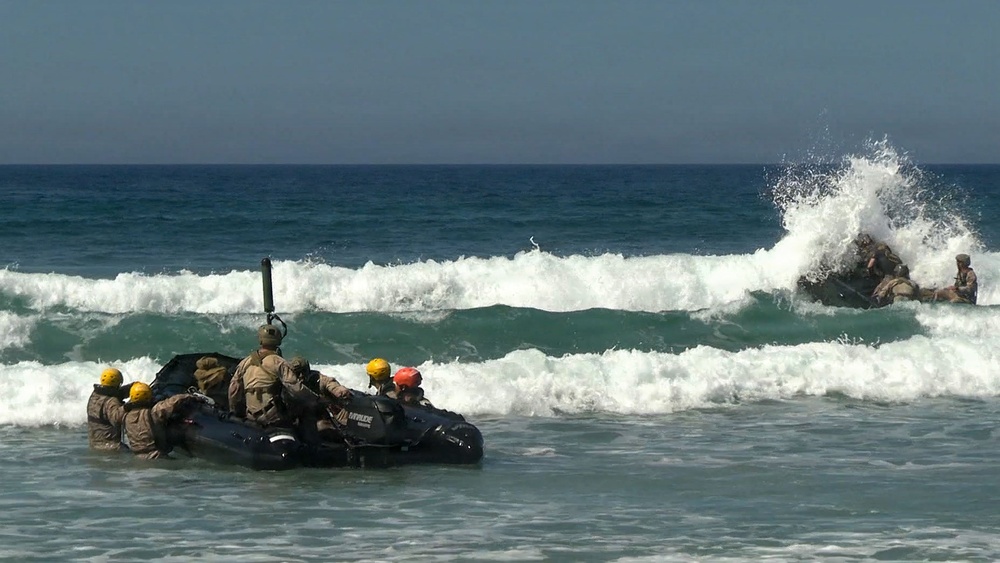 A group of military personnel in combat uniforms and yellow/orange helmets are navigating a black inflatable rubber raiding craft (IBS/Zodiac-style) through surf zone waves near a beach. A second group is visible in the background being engulfed by a breaking wave. This appears to be a military surf passage or amphibious training exercise, consistent with Navy SEAL BUD/S or similar special operations training.