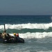 A group of military personnel in combat uniforms and yellow/orange helmets are navigating a black inflatable rubber raiding craft (IBS/Zodiac-style) through surf zone waves near a beach. A second group is visible in the background being engulfed by a breaking wave. This appears to be a military surf passage or amphibious training exercise, consistent with Navy SEAL BUD/S or similar special operations training.