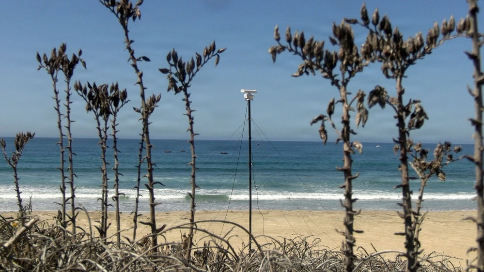 A coastal beach scene viewed through dried vegetation in the foreground, with a white camera or monitoring device mounted on a tripod with guy-wires visible in the center. The ocean stretches across the background with several small watercraft visible on the water. The sky is clear and blue with light haze near the horizon.