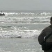 A military personnel in camouflage uniform stands on a beach shoreline observing what appears to be an amphibious assault vehicle (AAV) navigating through surf and waves in the ocean. The scene suggests a military amphibious training exercise or operation. The soldier is viewed from behind, facing the water where the vehicle is partially visible amid breaking waves.