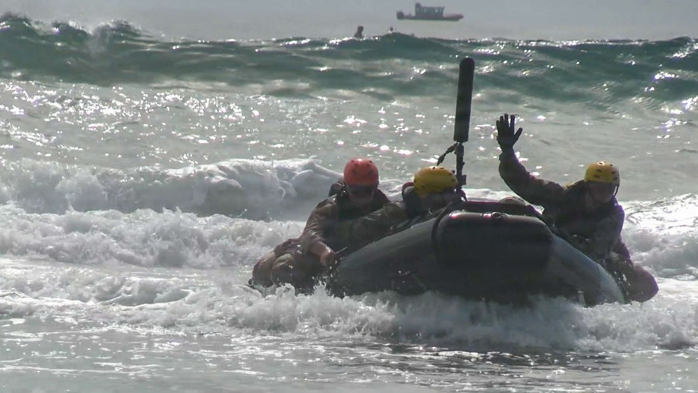A group of military or rescue personnel are navigating a rubber inflatable boat (IBS/Zodiac-type) through breaking surf near a shoreline. The individuals are wearing helmets and tactical/military-style clothing, suggesting this is a training exercise. One person has their hand raised, possibly signaling, while a larger vessel is visible in the background on the open water.