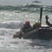 A group of military or rescue personnel are navigating a rubber inflatable boat (IBS/Zodiac-type) through breaking surf near a shoreline. The individuals are wearing helmets and tactical/military-style clothing, suggesting this is a training exercise. One person has their hand raised, possibly signaling, while a larger vessel is visible in the background on the open water.