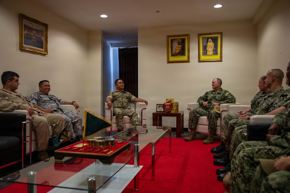 A formal military meeting is taking place in an official reception room, with personnel from what appear to be multiple nations seated in a circular arrangement. The setting includes framed portraits on the walls, a glass coffee table with ceremonial items, and a small flag, suggesting a diplomatic or bilateral military engagement. Personnel are wearing various military uniforms including US Navy digital camouflage, US Army combat uniforms, and what appears to be a host nation military uniform.