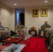 A formal military meeting is taking place in an official reception room, with personnel from what appear to be multiple nations seated in a circular arrangement. The setting includes framed portraits on the walls, a glass coffee table with ceremonial items, and a small flag, suggesting a diplomatic or bilateral military engagement. Personnel are wearing various military uniforms including US Navy digital camouflage, US Army combat uniforms, and what appears to be a host nation military uniform.