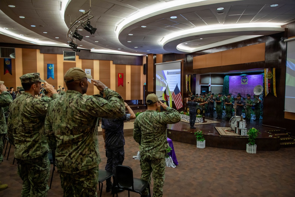 A formal military ceremony is taking place in an indoor auditorium or conference hall, where personnel in camouflage uniforms are rendering salutes toward a stage. On the stage, a military band is performing, with both an American flag and what appears to be a Bruneian flag visible, suggesting a multinational or bilateral military event. The setting and flags indicate this is likely a joint military exercise or official ceremony between the United States and Brunei.