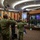 A formal military ceremony is taking place in an indoor auditorium or conference hall, where personnel in camouflage uniforms are rendering salutes toward a stage. On the stage, a military band is performing, with both an American flag and what appears to be a Bruneian flag visible, suggesting a multinational or bilateral military event. The setting and flags indicate this is likely a joint military exercise or official ceremony between the United States and Brunei.