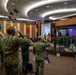 A formal military ceremony is taking place in an indoor auditorium or conference hall, where personnel in camouflage uniforms are rendering salutes toward a stage. On the stage, a military band is performing, with both an American flag and what appears to be a Bruneian flag visible, suggesting a multinational or bilateral military event. The setting and flags indicate this is likely a joint military exercise or official ceremony between the United States and Brunei.