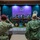Two military personnel are saluting during a formal ceremony in an indoor venue. One wears British-style MultiCam uniform with a maroon beret, and the other wears US Navy Working Uniform (NWU Type III) with a tan cap. A military band is performing on a decorated stage featuring traditional percussion instruments, with the US flag and what appears to be the Brunei flag visible in the background.