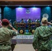 Two military personnel are saluting during a formal ceremony in an indoor venue. One wears British-style MultiCam uniform with a maroon beret, and the other wears US Navy Working Uniform (NWU Type III) with a tan cap. A military band is performing on a decorated stage featuring traditional percussion instruments, with the US flag and what appears to be the Brunei flag visible in the background.