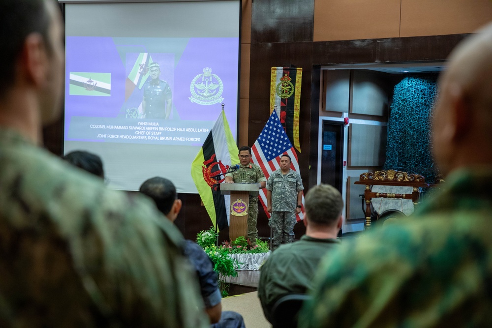A military ceremony or briefing is taking place in what appears to be a formal hall, with a uniformed officer speaking at a podium. The event involves personnel from multiple nations, as evidenced by the presence of both the United States flag and what appears to be the Brunei flag. A projection screen displays the name and title of Colonel Muhammad Suwardi Ariffin Bin PLDP Haji Abdul Latif, Chief of Staff, Joint Force Headquarters, Royal Brunei Armed Forces.