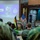 A military ceremony or briefing is taking place in what appears to be a formal hall, with a uniformed officer speaking at a podium. The event involves personnel from multiple nations, as evidenced by the presence of both the United States flag and what appears to be the Brunei flag. A projection screen displays the name and title of Colonel Muhammad Suwardi Ariffin Bin PLDP Haji Abdul Latif, Chief of Staff, Joint Force Headquarters, Royal Brunei Armed Forces.