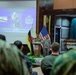 A military ceremony or briefing is taking place in what appears to be a formal hall, with a uniformed officer speaking at a podium. The event involves personnel from multiple nations, as evidenced by the presence of both the United States flag and what appears to be the Brunei flag. A projection screen displays the name and title of Colonel Muhammad Suwardi Ariffin Bin PLDP Haji Abdul Latif, Chief of Staff, Joint Force Headquarters, Royal Brunei Armed Forces.