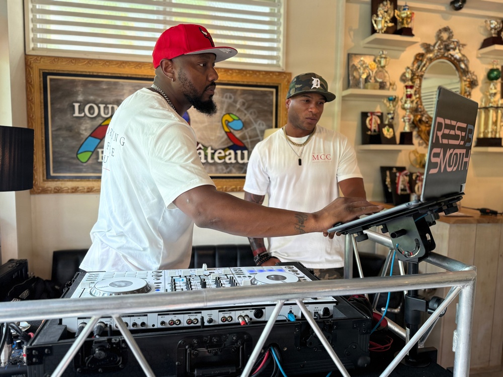 Two men are positioned behind a professional DJ setup in what appears to be an indoor venue or home studio. The man in the foreground wearing a red cap is actively operating the DJ controller and laptop, while the second man in a camouflage Detroit Tigers cap observes. The background features trophies on shelves, a framed sign reading 'Château,' and a laptop stand branded 'REESE SMOOTH'.