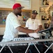 Two men are positioned behind a professional DJ setup in what appears to be an indoor venue or home studio. The man in the foreground wearing a red cap is actively operating the DJ controller and laptop, while the second man in a camouflage Detroit Tigers cap observes. The background features trophies on shelves, a framed sign reading 'Château,' and a laptop stand branded 'REESE SMOOTH'.