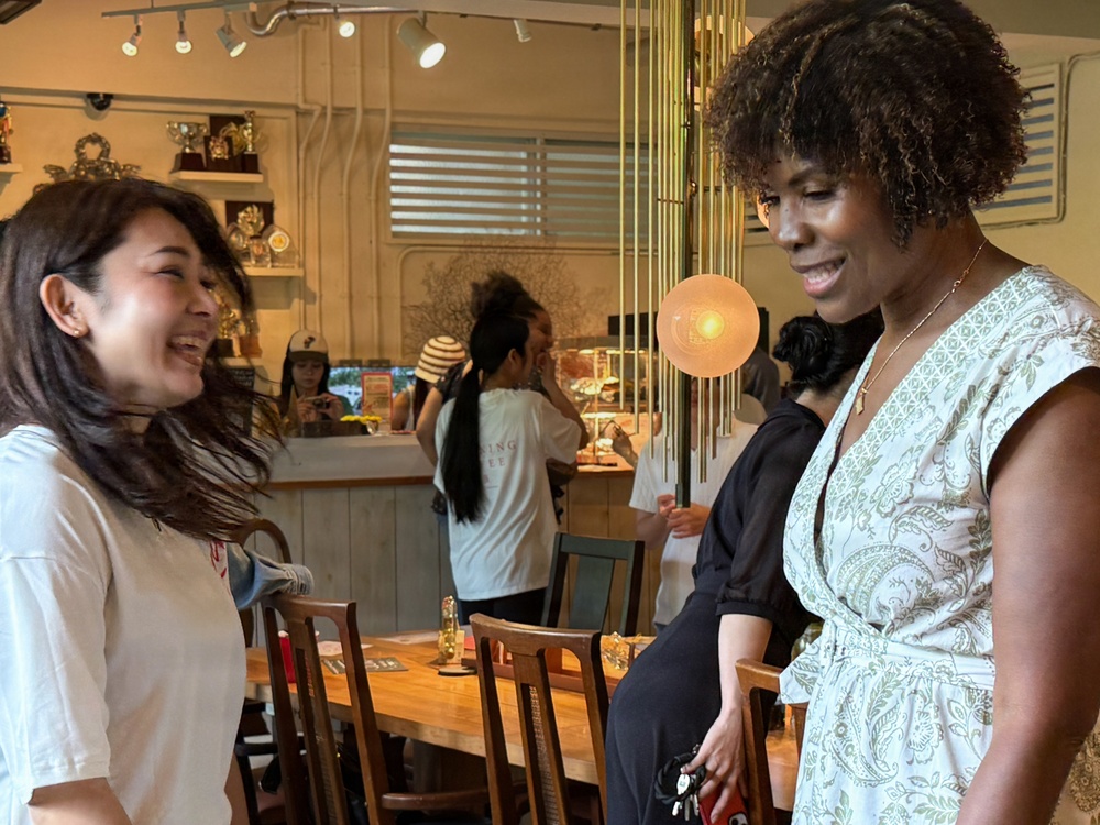 Two women are engaged in a cheerful conversation in what appears to be a café or restaurant setting. The woman on the left is laughing with her hair in motion, while the woman on the right in a floral wrap dress smiles warmly. The background shows other patrons and staff near a counter area with decorative shelving.