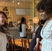 Two women are engaged in a cheerful conversation in what appears to be a café or restaurant setting. The woman on the left is laughing with her hair in motion, while the woman on the right in a floral wrap dress smiles warmly. The background shows other patrons and staff near a counter area with decorative shelving.