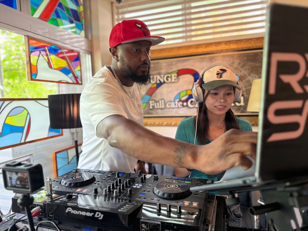 Two people are operating a Pioneer DJ controller setup inside what appears to be a café or lounge venue. A man wearing a red Chicago Bulls cap and white t-shirt with a silver chain is actively adjusting the DJ equipment, while a woman wearing a baseball cap and headphones stands beside him, observing or learning. The setting features colorful stained glass artwork on the walls and a sign partially reading 'Lounge' and 'Full cafe.'