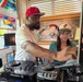 Two people are operating a Pioneer DJ controller setup inside what appears to be a café or lounge venue. A man wearing a red Chicago Bulls cap and white t-shirt with a silver chain is actively adjusting the DJ equipment, while a woman wearing a baseball cap and headphones stands beside him, observing or learning. The setting features colorful stained glass artwork on the walls and a sign partially reading 'Lounge' and 'Full cafe.'