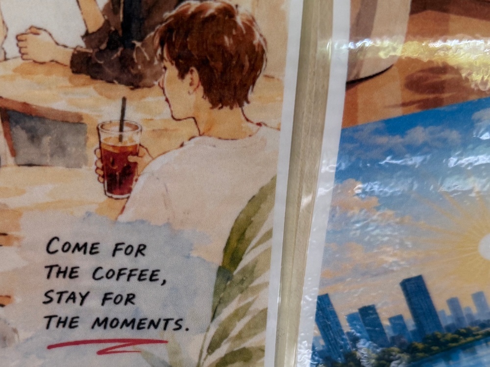 The image shows two printed artworks or posters placed side by side, partially wrapped in plastic. The left poster features a watercolor-style illustration of a person holding an iced coffee drink, accompanied by the text 'Come for the coffee, stay for the moments.' The right poster appears to depict a cityscape with blue skies and tall buildings.